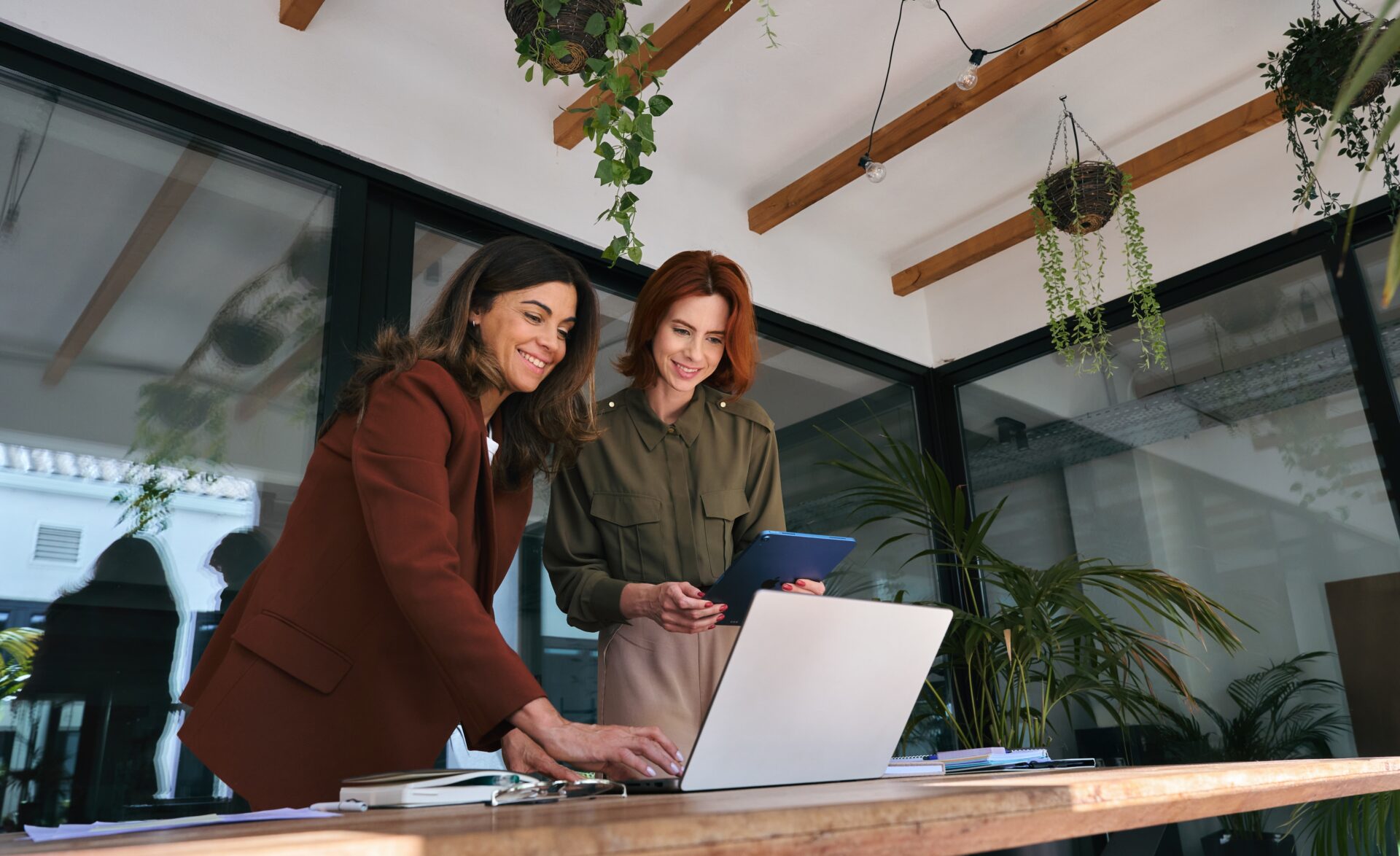 Two businesswomen smiling as they collaborate at a wooden table, one using a laptop while the other holds a tablet, in a bright modern office with large windows and hanging plants.