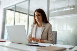 Professional woman in a modern office smiling while working on a laptop, representing focused, effective digital outreach and communication.