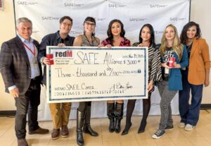 Seven people smile while holding an oversized ceremonial check for $3,000 from redM Austin to SAFE Alliance, in front of a SAFE "Stop Abuse for Everyone" backdrop.