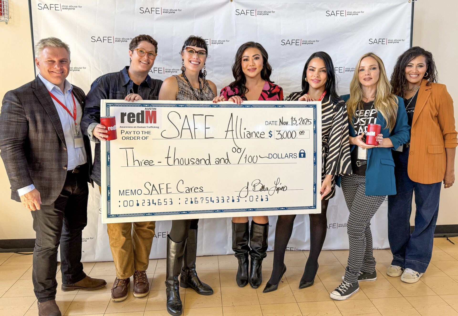Seven people smile while holding an oversized ceremonial check for $3,000 from redM Austin to SAFE Alliance, in front of a SAFE "Stop Abuse for Everyone" backdrop.