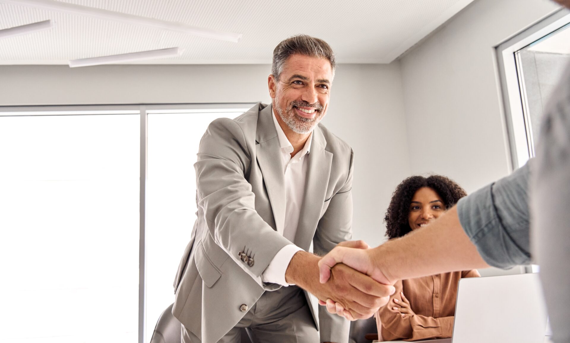 Happy older middle aged businessman leader shaking hand of new male partner, client or customer making sales deal at team executive board meeting in conference room. Business handshake concept.